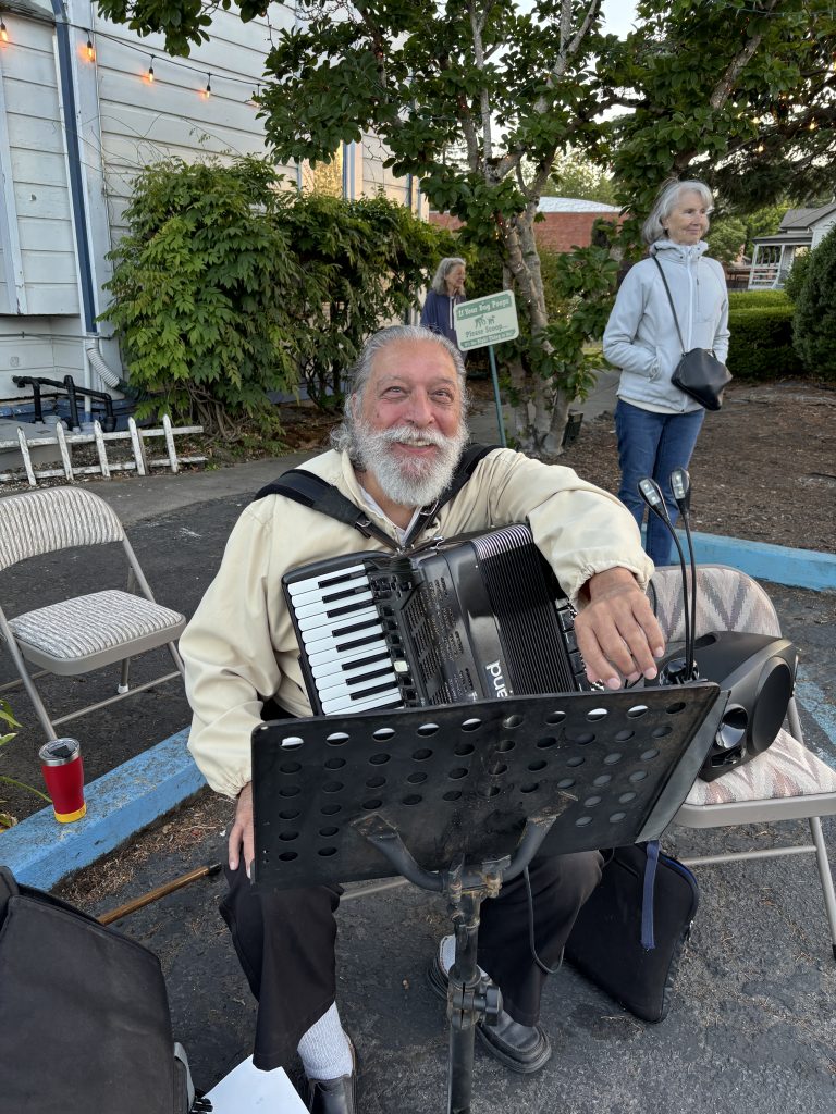 Accordionist Tony Mustaro smiles at the camera while seated in the parking lot at 620 Petaluma Blvd. North on June 14, 2026. Tony's wife is standing in the background. Photo taken by Katherine J. Rinehart